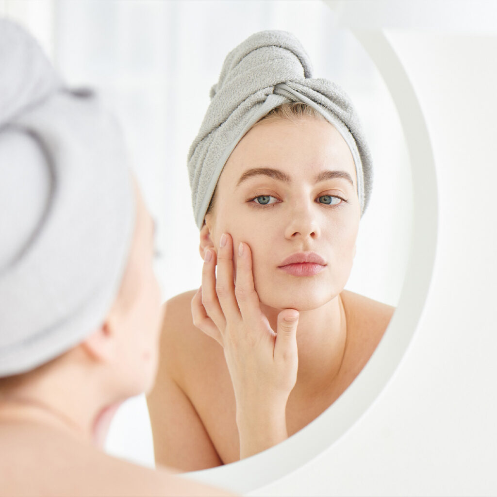 Photo of a woman with her hair wrapped in a towel checking out her skin in the bathroom mirror